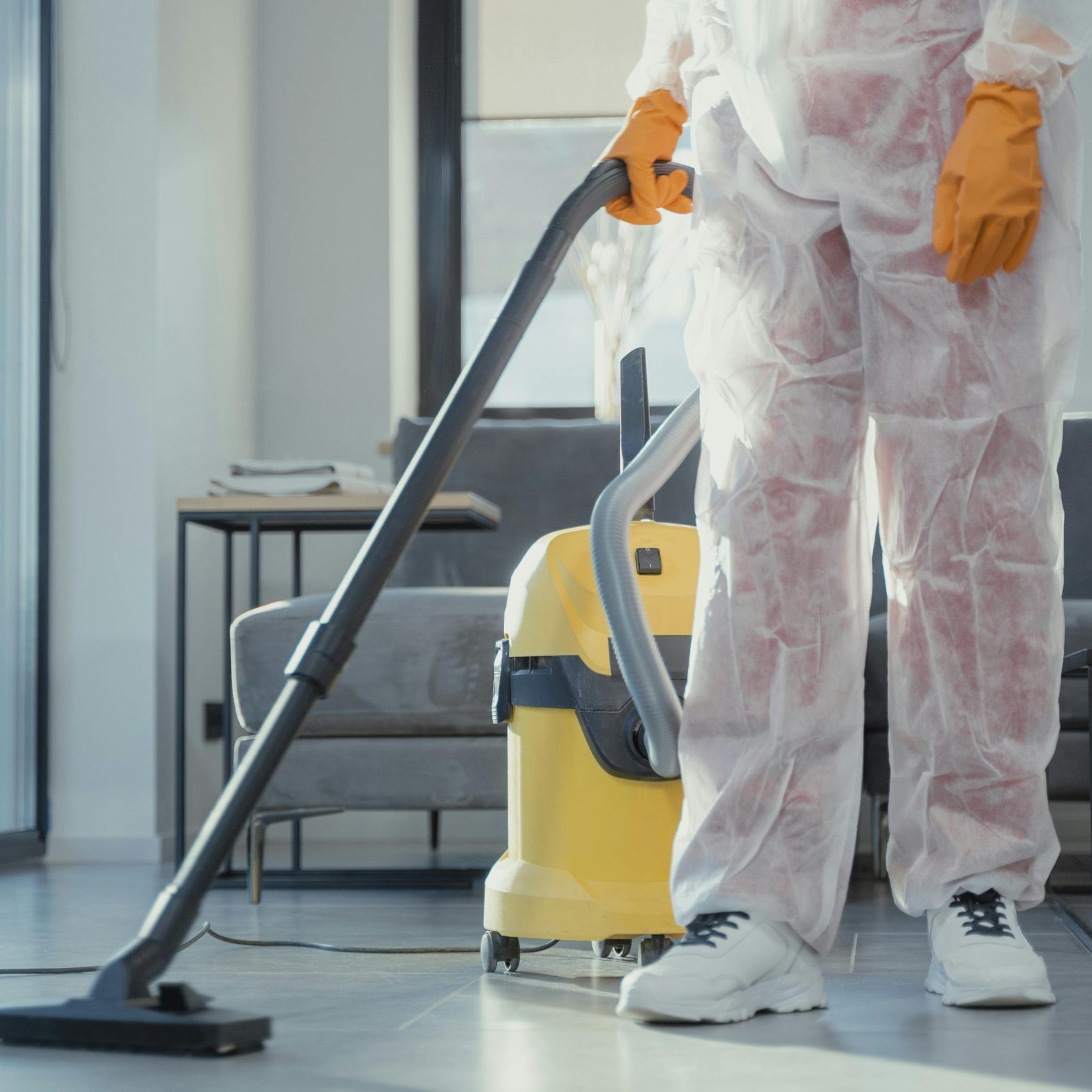 A cleaner in protective coveralls and gloves vacuuming a modern living room.