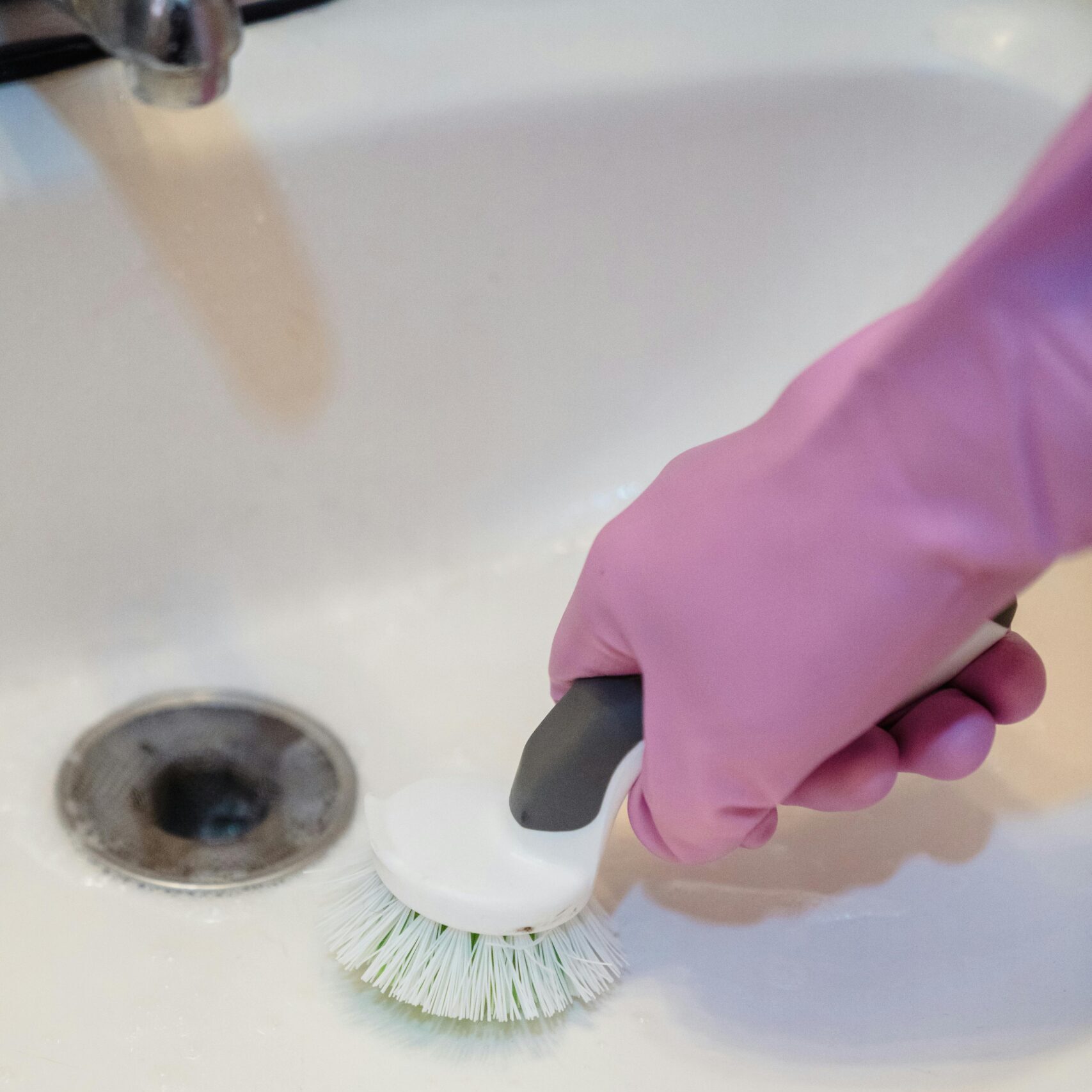 A close-up of a hand in a glove cleaning a sink with a scrub brush.